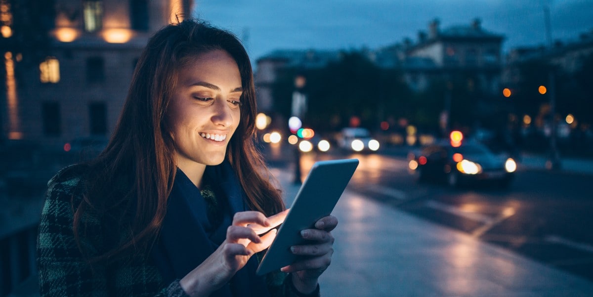 woman traveler using a tablet, waiting outside city building at twilight